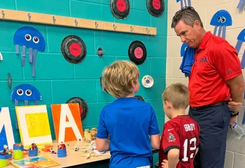 Man standing at craft display table with two children as they show off their work.